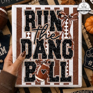 May include: A white rectangular graphic print featuring the text RUN THE DANG BALL in large black block letters. The design includes a brown football texture background with vertical white stripes, a brown football with a heart, and a brown bow.