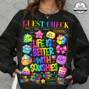 May include: A black long-sleeve sweatshirt featuring a colorful graphic of a guest check with the text "GUEST CHECK 019280" and "LIFE IS BETTER WITH SQUISHIES". The design includes various cartoon characters, a pink bow, and small hearts.