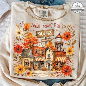 May include: A cream-colored sweatshirt featuring a graphic of a small town street with autumn foliage, orange and yellow flowers, a water tower, and a wooden sign reading Small town Fall 90048. The design includes brick buildings with awnings.