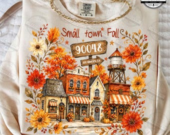 May include: A cream-colored sweatshirt featuring a graphic of a small town street with autumn foliage, orange and yellow flowers, a water tower, and a wooden sign reading Small town Fall 90048. The design includes brick buildings with awnings.