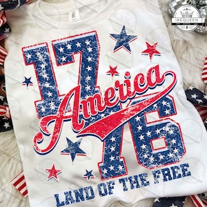 Könnte beinhalten: Weißes T-Shirt mit einem Used-Design mit den Zahlen "1776" in Blau und Rot, gefüllt mit einem Sternmuster. Das Wort "America" ist in roter Schrift geschrieben, und der Satz "Land of the Free" steht unten. Patriotisches Thema.