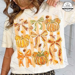 Puede incluir: Una camiseta de color crema con un estampado gráfico de calabazas naranjas con orejas de ratón y varios lazos decorativos en tonos naranja, amarillo y verde. El diseño incluye pequeñas hojas de otoño y el texto IREQUEEN en la parte superior.