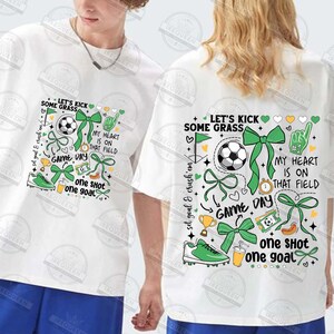 May include: White t-shirt with a green, white, and yellow graphic design featuring a soccer ball, bows, hearts, and the text "Let's Kick Some Grass", "My Heart Is On That Field", "Game Day", and "One Shot One Goal".