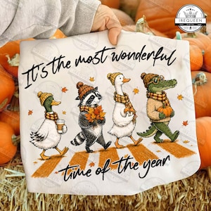 May include: A white t-shirt featuring a graphic of a duck, a raccoon, a goose, and an alligator walking in a line. They wear autumn-themed hats and scarves. The text reads, It's the most wonderful time of the year, with orange leaves scattered.