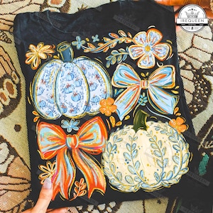 May include: Black t-shirt featuring a colorful fall design. The design includes pumpkins, bows, and floral elements in shades of blue, orange, and yellow. The pumpkins have intricate patterns, and the bows have a watercolor effect.