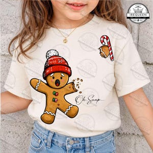 May include: A cream-colored short-sleeve t-shirt featuring a graphic of a gingerbread person wearing a red knit hat with white snowflakes. The gingerbread person has a missing arm, a surprised expression, and the text Oh Snap printed nearby.