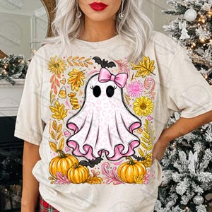 Może przedstawiać: Kremowy t-shirt z centralną grafiką przedstawiającą białego ducha w r&oacute;żowy wz&oacute;r panterki, noszącego r&oacute;żową kokardę. Duch jest otoczony ż&oacute;łtymi słonecznikami, pomarańczowymi dyniami, jesiennymi liśćmi, cukierkami i małymi czarnymi nietoperzami.