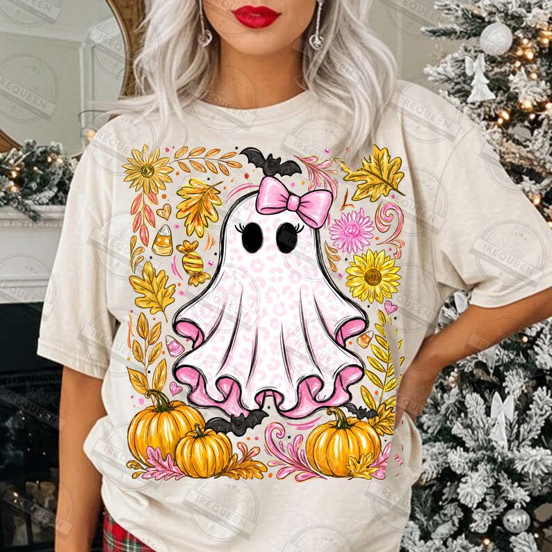Może przedstawiać: Kremowy t-shirt z centralną grafiką przedstawiającą białego ducha w r&oacute;żowy wz&oacute;r panterki, noszącego r&oacute;żową kokardę. Duch jest otoczony ż&oacute;łtymi słonecznikami, pomarańczowymi dyniami, jesiennymi liśćmi, cukierkami i małymi czarnymi nietoperzami.