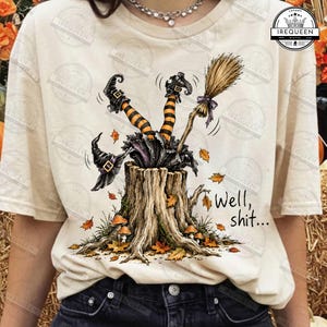May include: A cream-colored t-shirt featuring a graphic of a tree stump with a witch's legs in orange and black striped stockings sticking out of the top. A broom and hat are nearby, surrounded by autumn leaves and the text "Well, shit..."