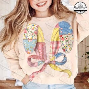 Peut inclure: Un sweat-shirt beige orné d'un motif d'oreilles de lapin en patchwork. Les oreilles sont faites de tissus à motifs floraux et vichy aux couleurs pastel, avec un nœud rose à la base. Le motif est cousu sur le sweat-shirt.