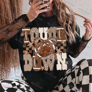 Peut inclure: Un t-shirt noir avec le texte "TOUCH DOWN" en grandes lettres majuscules aux motifs bruns et blancs vari&eacute;s, incluant des imprim&eacute;s l&eacute;opard, carreaux et rayures. Un ballon de football brun avec un grand n&oelig;ud est centr&eacute; sur le motif.