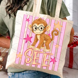 Puede incluir: Un bolso tote beige con un mono de dibujos animados con gafas, sobre un fondo de rayas rosas con estrellas. La palabra "SEE" se muestra en una fuente l&uacute;dica debajo del mono. El bolso lo sostiene una persona.