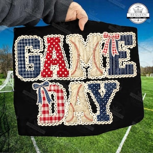 Puede incluir: Camiseta negra con las palabras "GAME DAY" en un diseño de patchwork. Las letras presentan varios patrones, incluyendo cuadros, lunares y costuras de béisbol, con lazos decorativos y un borde festoneado.