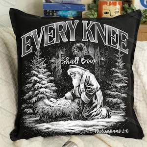 May include: Black decorative pillow with a Christmas scene. The pillow features a black and white illustration of Santa Claus kneeling before baby Jesus. The text "EVERY KNEE SHALL BOW" is at the top, with "Philippians 2:10" at the bottom.
