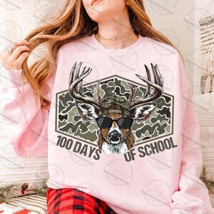 Puede incluir: Sudadera rosa claro con un dise&ntilde;o de cabeza de ciervo con gafas de sol y un sombrero de camuflaje, sobre un fondo de camuflaje hexagonal. El texto "100 DAYS OF SCHOOL" se muestra debajo.
