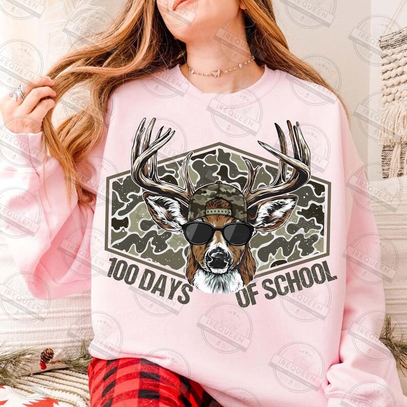 Puede incluir: Sudadera rosa claro con un dise&ntilde;o de cabeza de ciervo con gafas de sol y un sombrero de camuflaje, sobre un fondo de camuflaje hexagonal. El texto "100 DAYS OF SCHOOL" se muestra debajo.