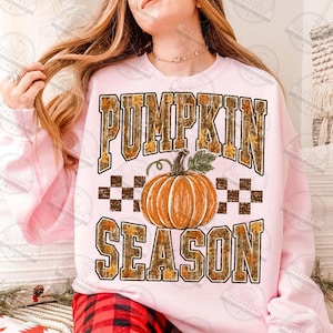 May include: A light pink sweatshirt featuring the text PUMPKIN SEASON in large block letters with a camouflage and autumn leaf pattern. Centered between the words is a graphic of a bright orange pumpkin with a green stem and leaves.