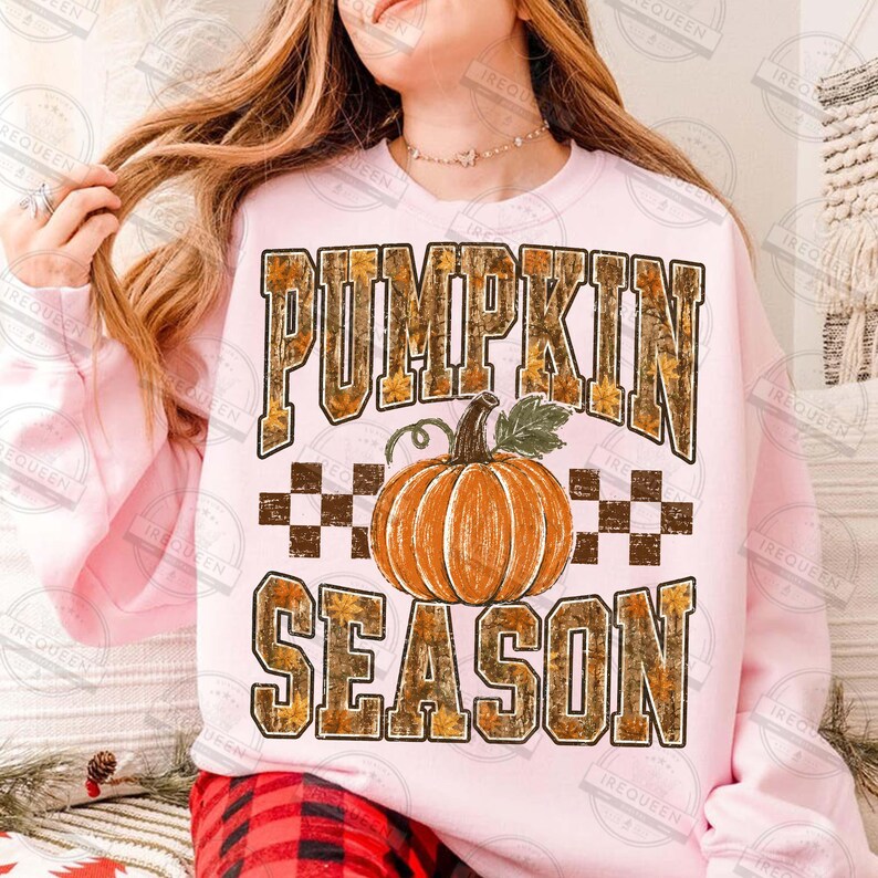 May include: A light pink sweatshirt featuring the text PUMPKIN SEASON in large block letters with a camouflage and autumn leaf pattern. Centered between the words is a graphic of a bright orange pumpkin with a green stem and leaves.