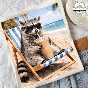May include: A graphic tee featuring a raccoon wearing sunglasses, relaxing in a beach chair, and drinking an iced coffee. The raccoon is on a beach with blue water and palm trees in the background. The tee is on a denim surface.