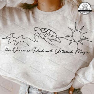 May include: Pale grey sweatshirt featuring a black line drawing of waves, a turtle, and a sun. The words "The Ocean is Filled with Untamed Magic" are below the drawing. A circular logo is in the upper right corner.