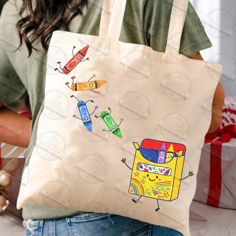 May include: A beige canvas tote bag featuring a graphic print of a yellow crayon box with arms and legs, surrounded by four floating, animated crayons in red, orange, blue, and green. The yellow box contains two additional crayons and displays colourful scribbles.