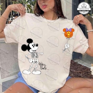 以下が含まれることがあります： クリーム色のTシャツで、スケルトンのネズミのキャラクターの白黒グラフィックが描かれています。キャラクターは、紫色の水玉模様の蝶ネクタイが付いたオレンジ色のカボチャの風船につながった紐を持っています。黒色で「Well, Shit...」という文字がプリントされています。
