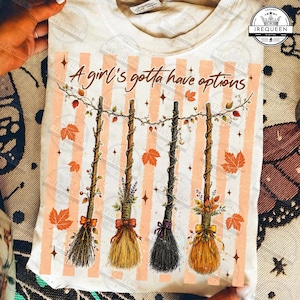 May include: A cream-colored t-shirt featuring a graphic of four hanging broomsticks decorated with autumn leaves and ribbons. The text "A girl's gotta have options" is printed above the brooms, set against a background of vertical peach stripes.