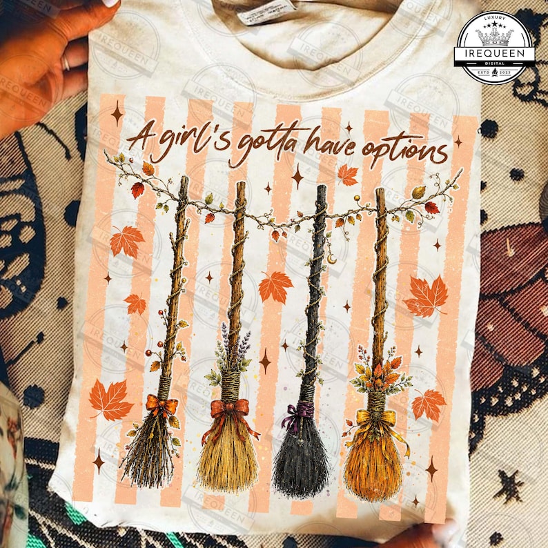 May include: A cream-colored t-shirt featuring a graphic of four hanging broomsticks decorated with autumn leaves and ribbons. The text "A girl's gotta have options" is printed above the brooms, set against a background of vertical peach stripes.