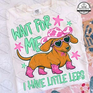 Op de afbeelding: Een crèmekleurig t-shirt met een bruine teckel die een roze cowboyhoed met koeienprint, hartvormige zonnebril en roze cowboylaarzen draagt. De tekst "WAIT FOR ME I HAVE LITTLE LEGS" staat in het groen, omringd door roze en groene sterren.