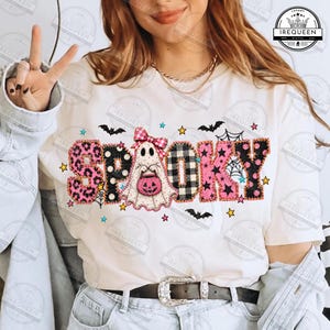 May include: A cream-colored t-shirt featuring the word SPOOKY in large, patterned letters. The letters include pink leopard print, black polka dots, black and white buffalo plaid, and pink stars. A ghost with a pink bow holds a pumpkin.