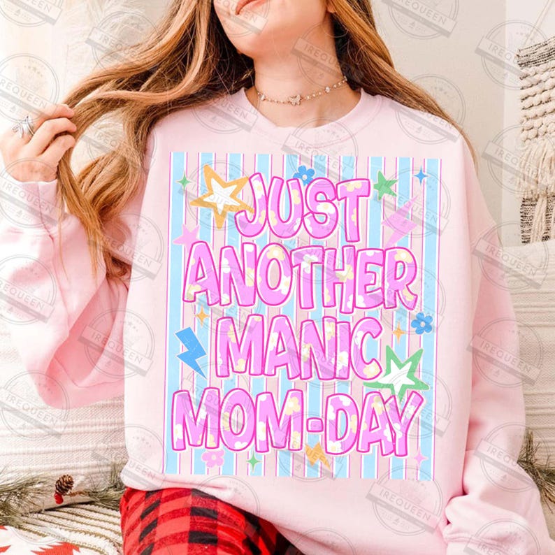 May include: A light pink sweatshirt with the text "JUST ANOTHER MANIC MOM-DAY" in pink, blue, and yellow. The text is set against a background of blue and pink stripes, with star and lightning bolt graphics.