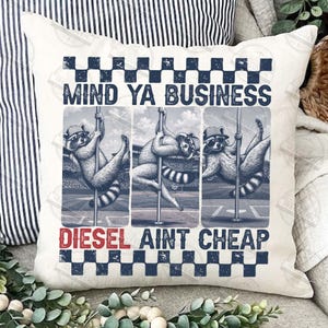 May include: A white pillow with a humorous graphic of raccoons pole dancing. The text "MIND YA BUSINESS" is above the raccoons, and "DIESEL AINT CHEAP" is below. The pillow has a checkered border.