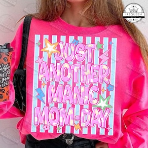 May include: A bright pink sweatshirt with the text "JUST ANOTHER MANIC MOM-DAY" in a playful, colorful font. The design includes stars, flowers, and lightning bolts. The sweatshirt has a crew neck and long sleeves.