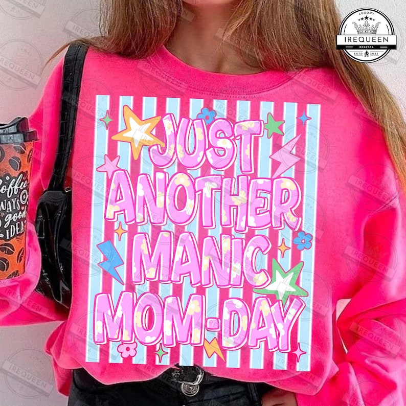 May include: A bright pink sweatshirt with the text "JUST ANOTHER MANIC MOM-DAY" in a playful, colorful font. The design includes stars, flowers, and lightning bolts. The sweatshirt has a crew neck and long sleeves.