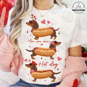 May include: White t-shirt with a fun design of three dachshunds in hot dog costumes, decorated with hearts and stars. The text "You are one Hot dog" is printed on the shirt. A lighthearted and quirky design.