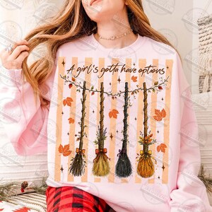May include: A light pink sweatshirt featuring a graphic of four hanging brooms decorated with autumn leaves and flowers. The text A girl's gotta have options is printed above the brooms, set against a background of vertical orange stripes.