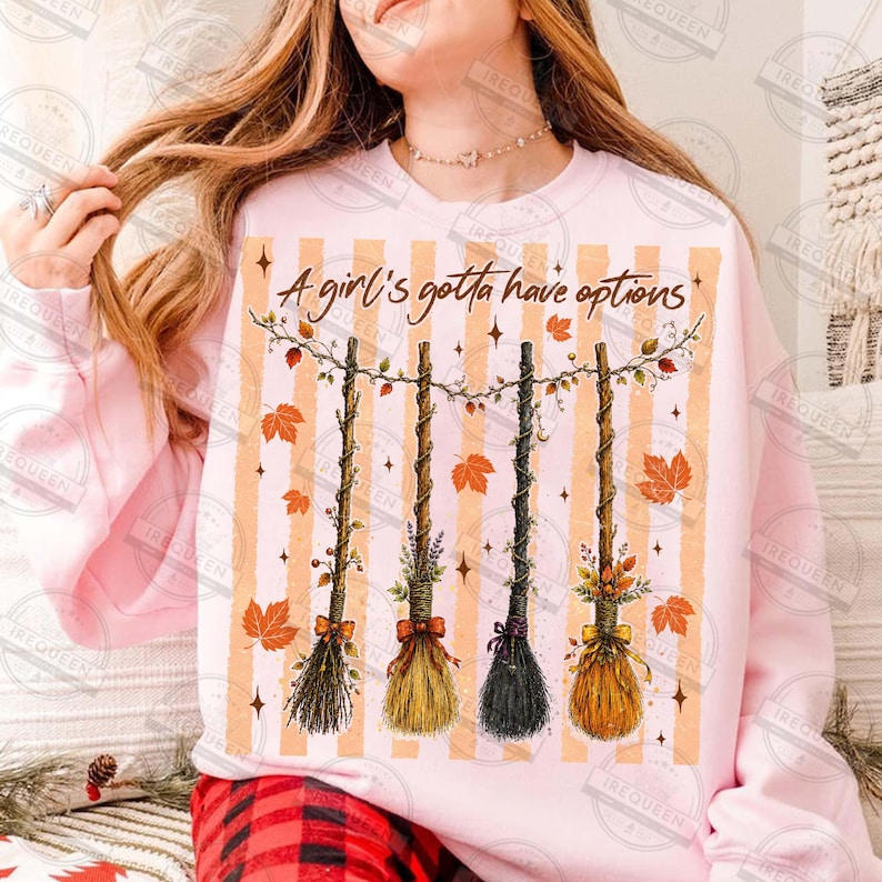 May include: A light pink sweatshirt featuring a graphic of four hanging brooms decorated with autumn leaves and flowers. The text A girl's gotta have options is printed above the brooms, set against a background of vertical orange stripes.