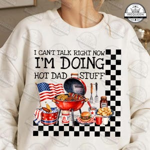 May include: White sweatshirt with a graphic of a red grill, American flag, and text that reads "I CAN'T TALK RIGHT NOW I'M DOING HOT DAD STUFF." The design includes a checkered pattern and BBQ-themed illustrations.