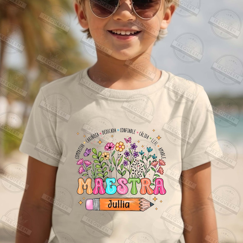 May include: A light beige t-shirt with a colorful graphic of flowers and a pencil with the word "MAESTRA" and the name "Jullia" written on it. The graphic is surrounded by words in Spanish describing positive qualities.
