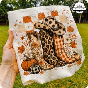 Puede incluir: Una camiseta blanca con un estampado gráfico de tres botas vaqueras, una con estampado floral, otra con estampado de leopardo y otra con estampado de cuadros, junto a un sombrero de vaquero, calabazas y hojas de otoño.