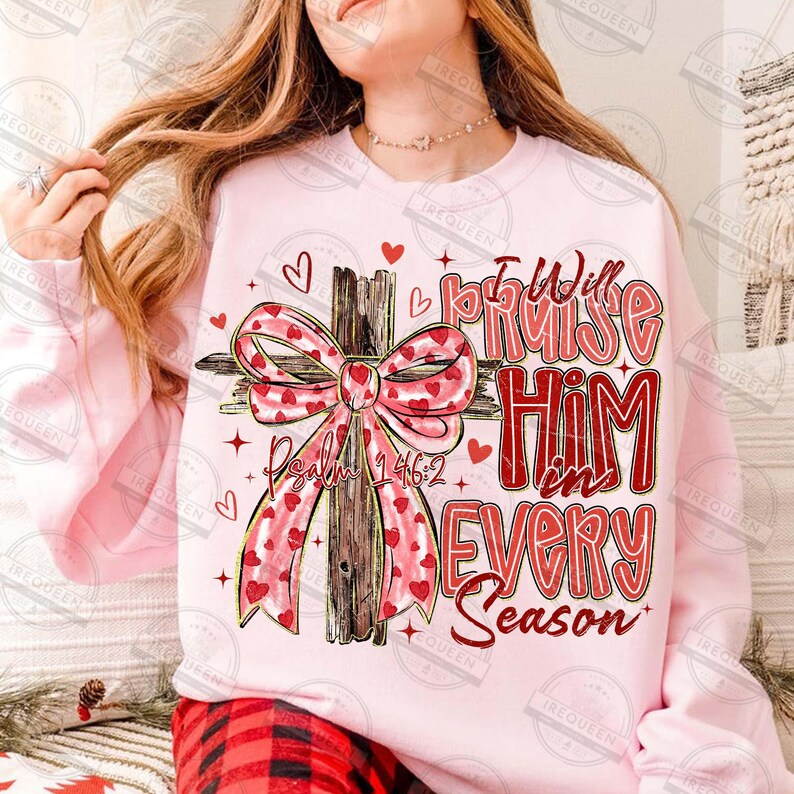 May include: A light pink sweatshirt with a cross design. The cross is adorned with a large bow featuring red hearts. The text reads "I Will Praise Him in Every Season" and "Psalm 146:2" in red.