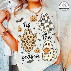 May include: A cream-colored t-shirt featuring a graphic print with the text 'tis the season'. The design includes three ghosts with different patterns, including checkered, leopard print, and polka dots, alongside small orange pumpkins and bats.