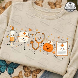 May include: A beige crewneck sweatshirt featuring a graphic print of five animated medical items: a syringe, a nurse cap, a stethoscope, a pumpkin wearing a nurse cap, and a clipboard, all with smiling faces and orange accents.