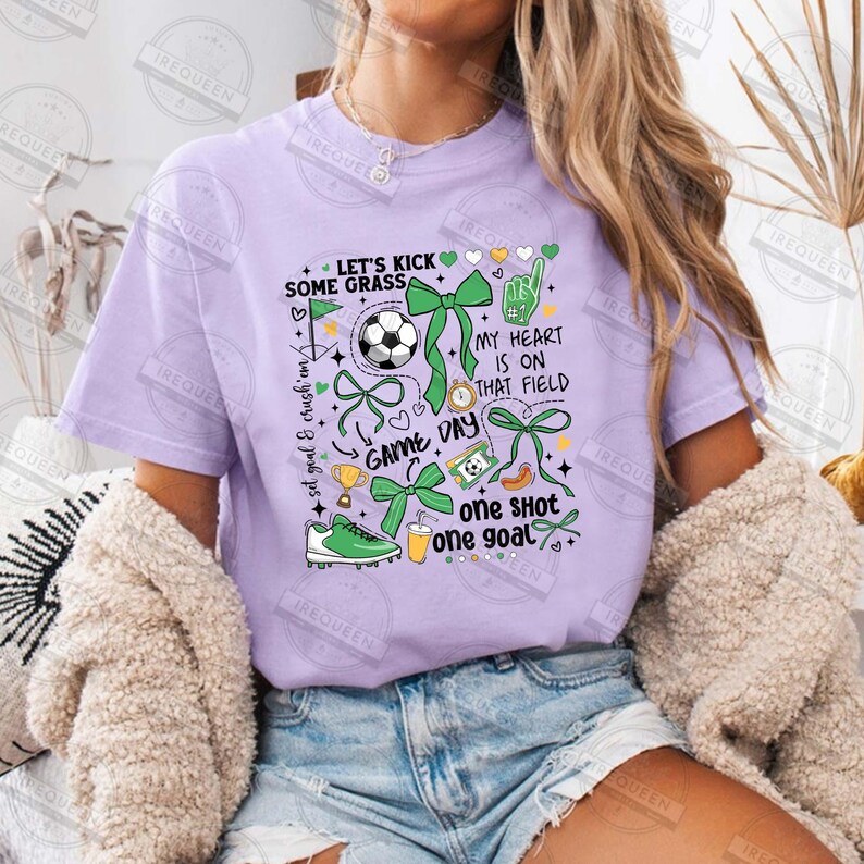 May include: Purple t-shirt with a green and yellow graphic design featuring soccer-themed illustrations, text, and hearts. The text includes "Let's Kick Some Grass", "Game Day", "My Heart Is On That Field", "One Shot One Goal", and "Set Goal & Crush 'Em".
