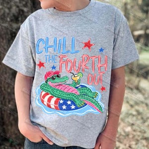May include: A grey t-shirt with a cartoon alligator wearing sunglasses and a hat, relaxing on a red, white, and blue float with a drink. The text "Chill the Fourth Out" is in blue and red, with star accents.