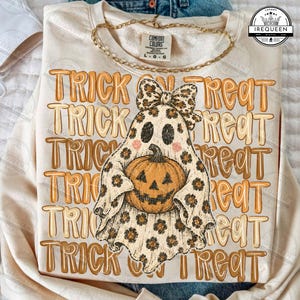 May include: A cream-colored sweatshirt featuring a repeating orange and brown text pattern that reads TRICK OR TREAT. Centered is a ghost illustration wearing a leopard print bow and holding a jack-o'-lantern pumpkin with a matching print.