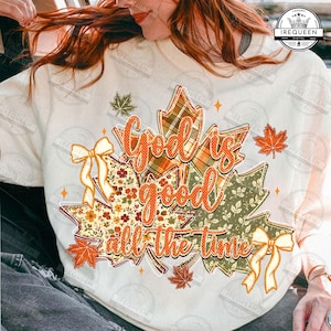 Peut inclure: Un sweat-shirt crème orné d'un motif de trois grandes feuilles d'érable aux motifs écossais, floraux et vert uni. Le texte "God is good all the time" est imprimé en orange sur les feuilles, agrémenté de deux rubans jaunes et de petites étoiles orange.
