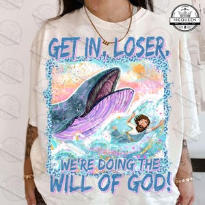 Puede incluir: Camiseta blanca con un diseño gráfico colorido de una ballena y una persona en el agua. El texto "GET IN, LOSER. WE'RE DOING THE WILL OF GOD!" está impreso en azul y rosa. El diseño tiene un borde con estampado de leopardo azul.