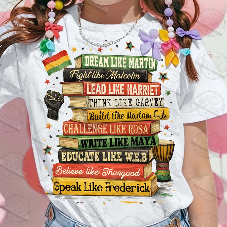 May include: White t-shirt featuring a stack of books with inspirational phrases like "Dream Like Martin" and "Fight Like Malcolm." The design includes a black power fist, a small flag, and a drum. The shirt is adorned with star accents.