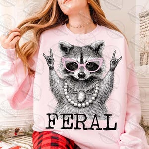 Puede incluir: Sudadera rosa claro con un mapache con gafas rosas y un collar de perlas. El mapache hace un gesto de rock and roll con la mano. La palabra "FERAL" est&aacute; impresa en letras negras.